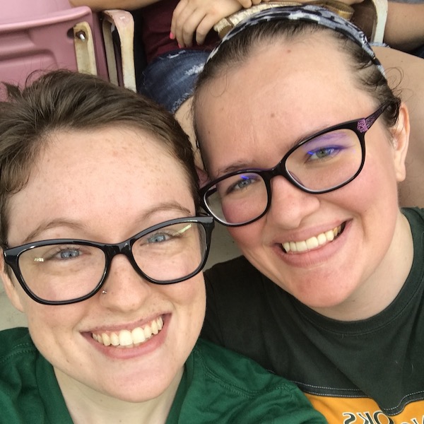 A white genderqueer person and a white woman in a sports stadium, heads pressed together